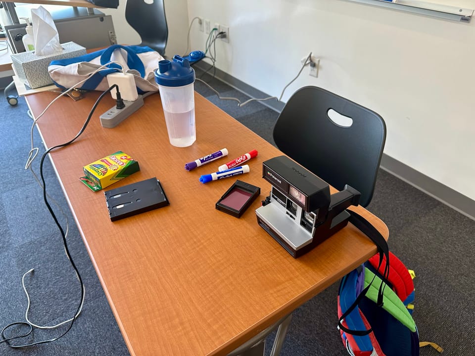 table with extension cord, polaroid camera, crayons, dry erase markers, and a water bottle